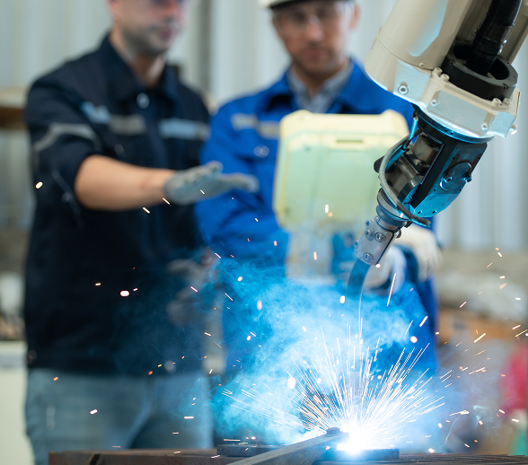 Brazo robótico realizando soldadura automatizada en una fábrica, con operarios supervisando el proceso en el fondo.