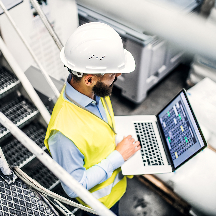 Supervisor industrial con casco y chaleco de seguridad monitoreando procesos en su laptop dentro de una planta de producción.